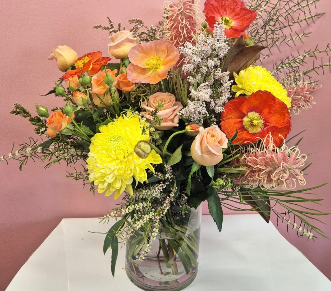 A vibrant, multicolor floral arrangement featuring yellow chrysanthemums, orange poppies, peach roses, and pink wildflowers in a clear glass vase, set against a dusty pink wall and white table surface.