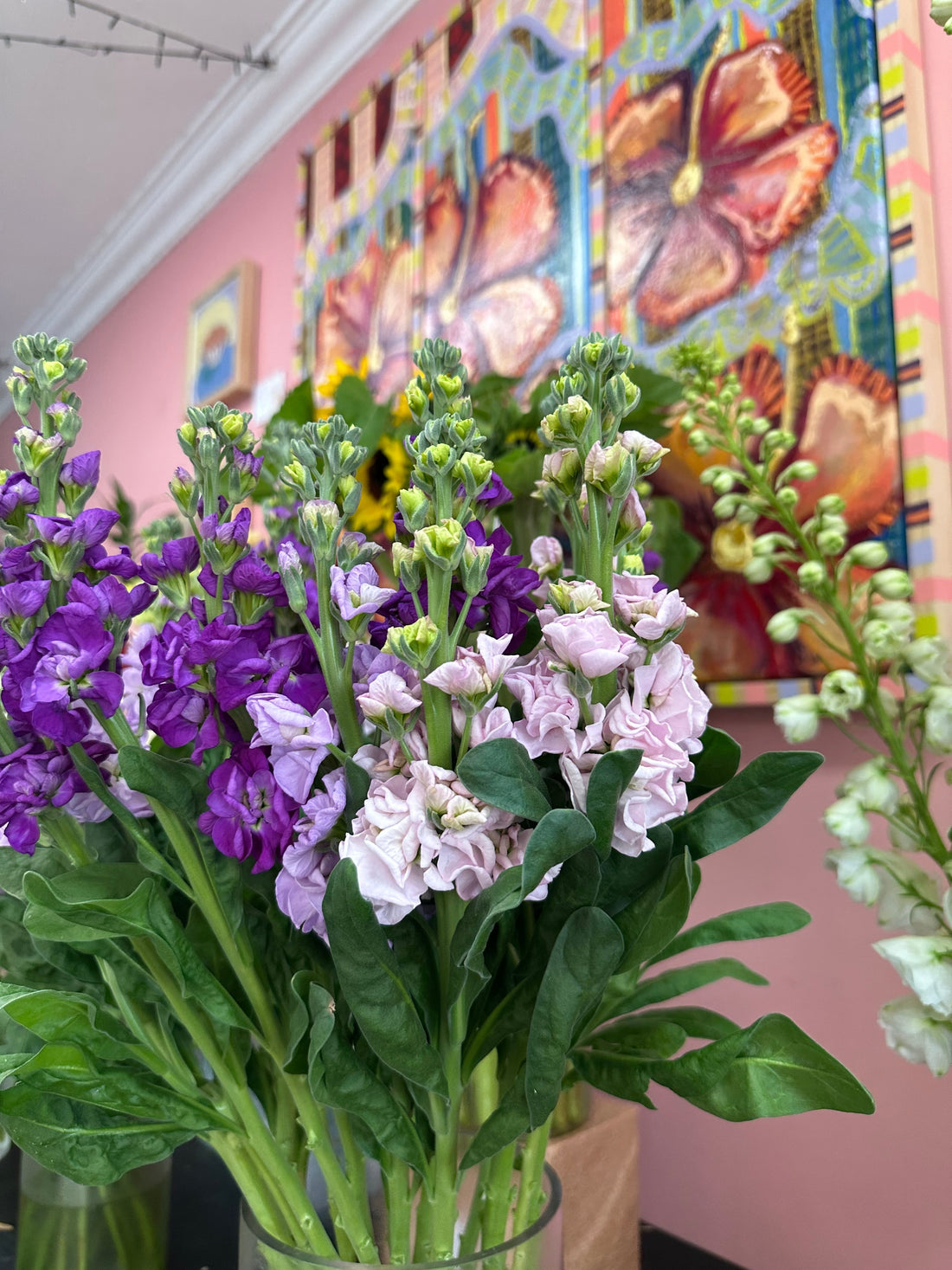 Purple and lavender flowers with hibiscus painting on pink wall behind flowers
