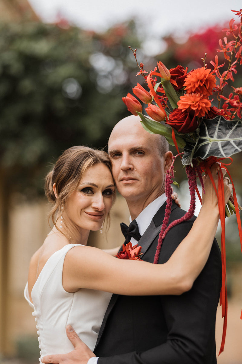 Couple in wedding attire with a modern bouquet of red flowers at Jimbour House.