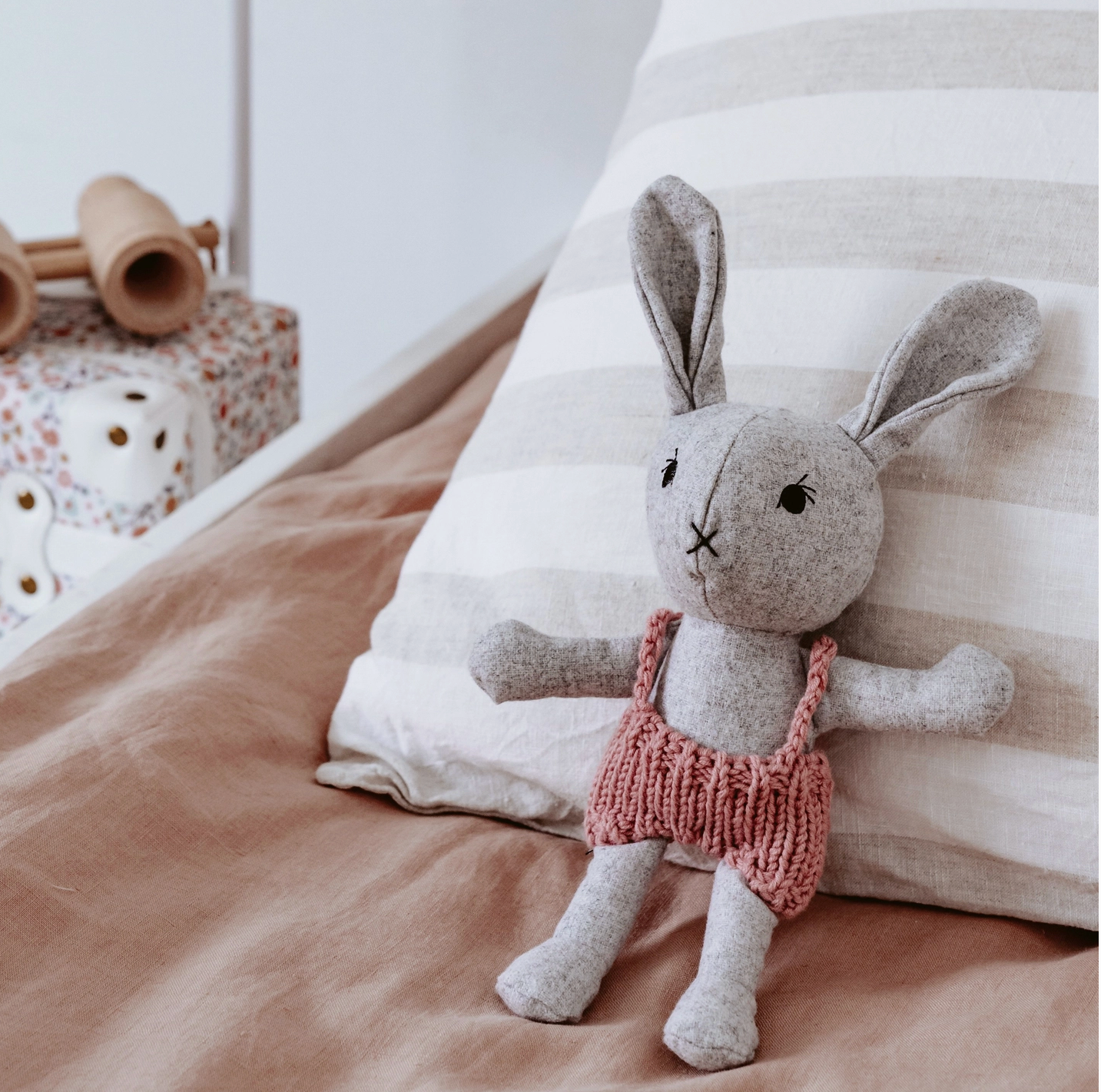 Stuffed rabbit toy with a pink outfit on a bed with striped pillow and brown blanket.