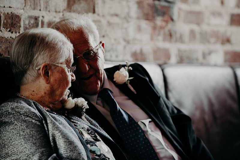 Two elderly people sitting close together with flowers on the suits against a brick wall.