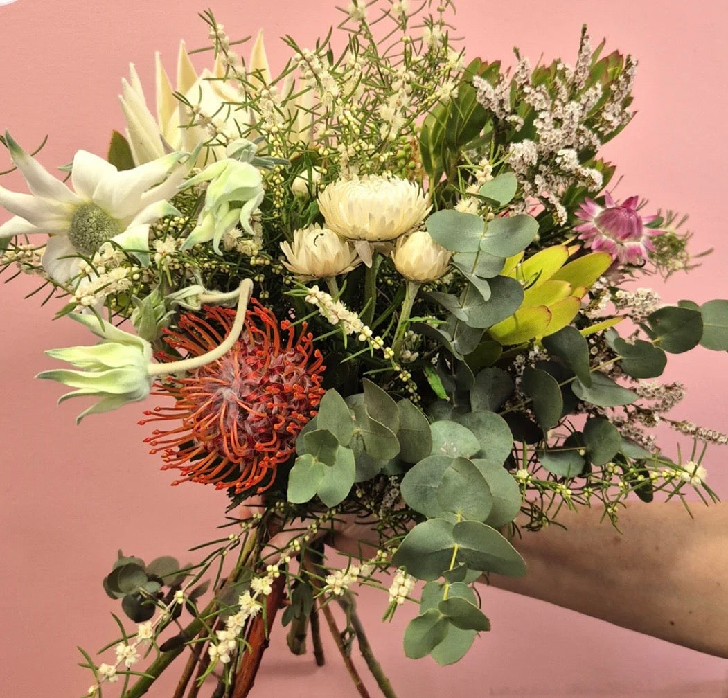 A hand holding a lush, mixed floral bouquet featuring white protea-like flowers, a prominent orange-red spiky flower, green eucalyptus leaves, and small white filler blooms against a solid dusty pink background.