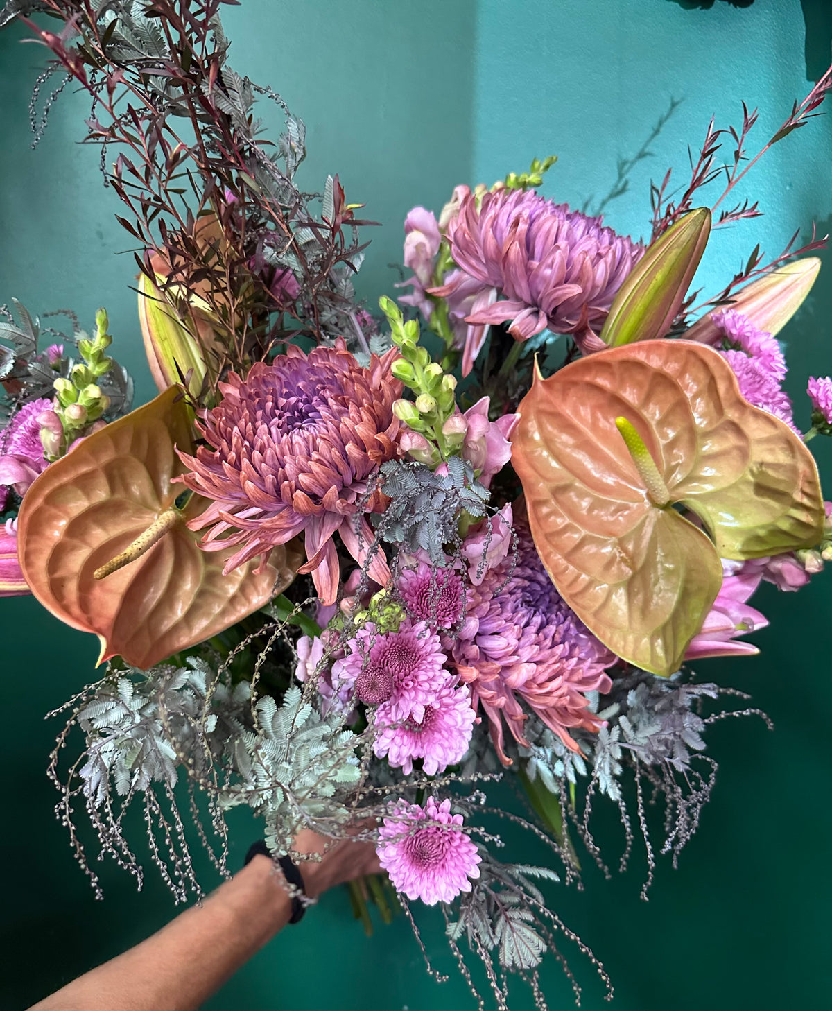 A vibrant artificial flower bouquet featuring pink chrysanthemums, a pink protea, bronze anthuriums, and dusty purple foliage, held by a person's hand against a teal background.