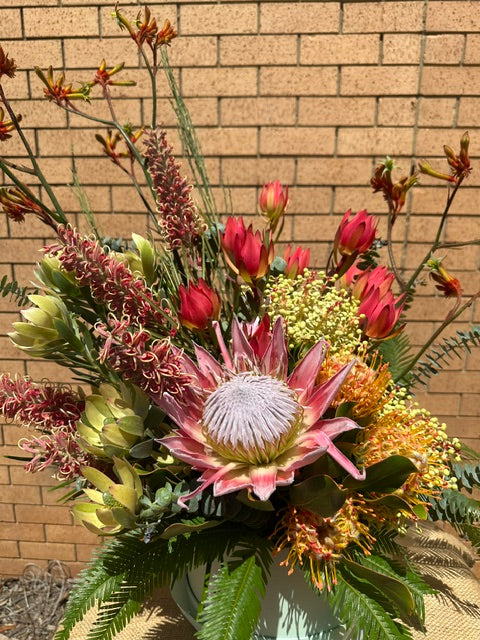 A vibrant artificial flower arrangement featuring a large pink and white protea, red waratahs, yellow gypsophila, and green ferns, set against a tan brick wall background.