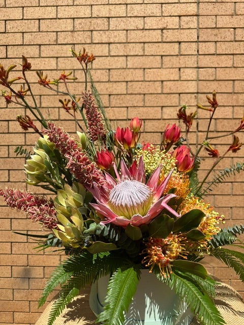 A vibrant floral arrangement featuring a prominent pink and white protea, red tulips, yellow flowers, and green foliage, set in a light blue-green pot against a tan brick wall background.