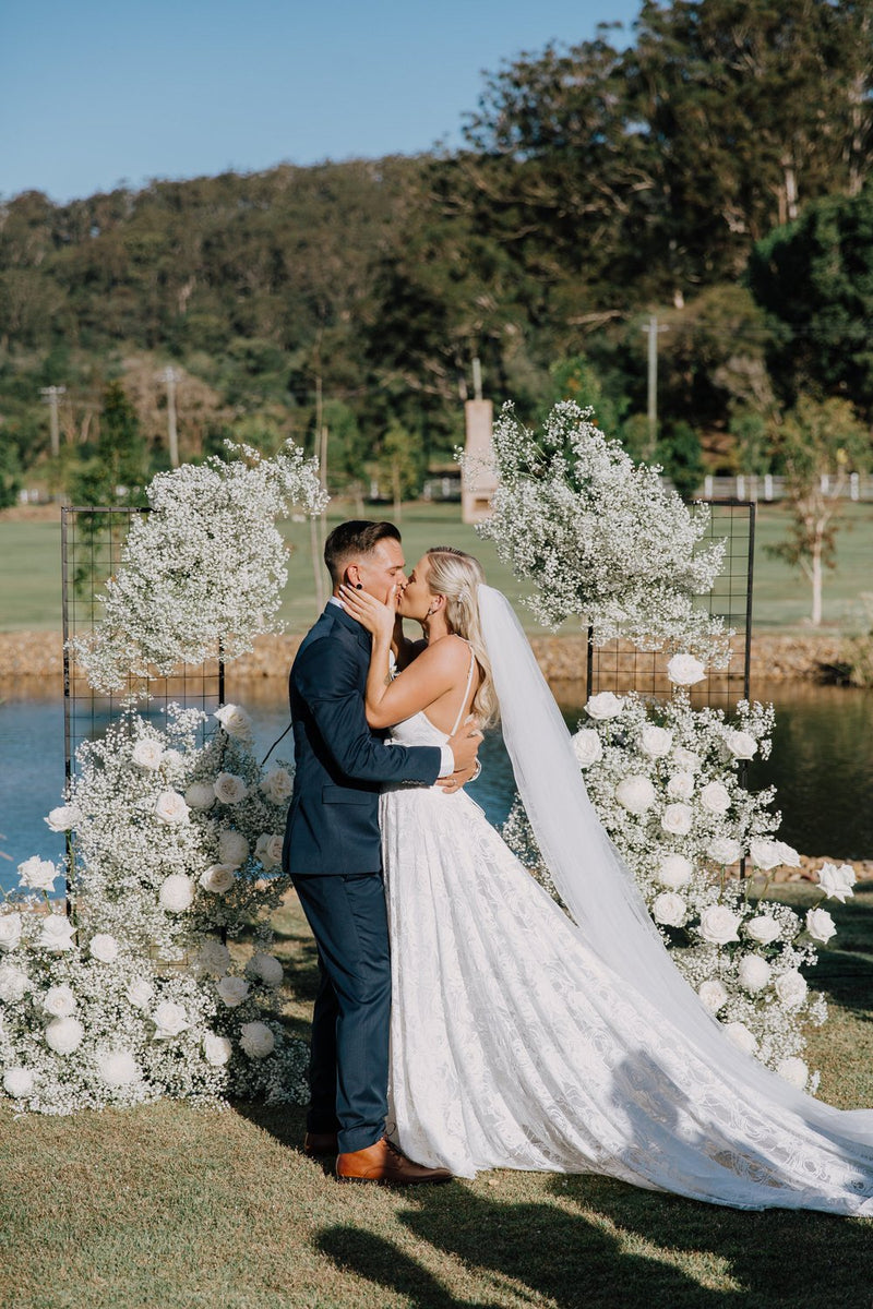Couple in wedding attire embracing in front of a gypsophila and white rose arch by a lake.