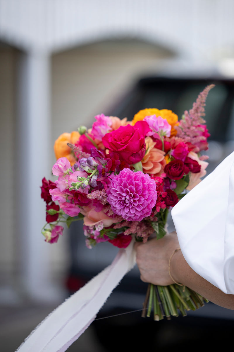 Bouquet of colorful flowers like dahlias, roses and sweet peas held by a person wearing a wedding dress.
