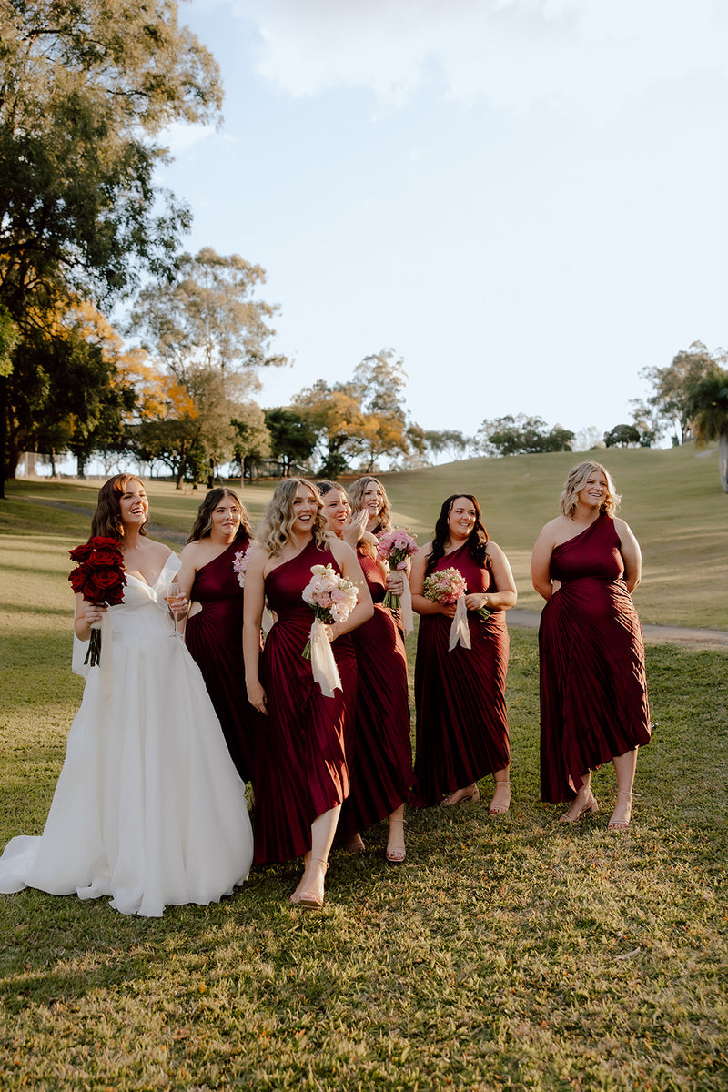 Bride and bridesmaids in burgundy dresses standing on a grassy field with trees in the background. With bridal bouquet in red roses. 