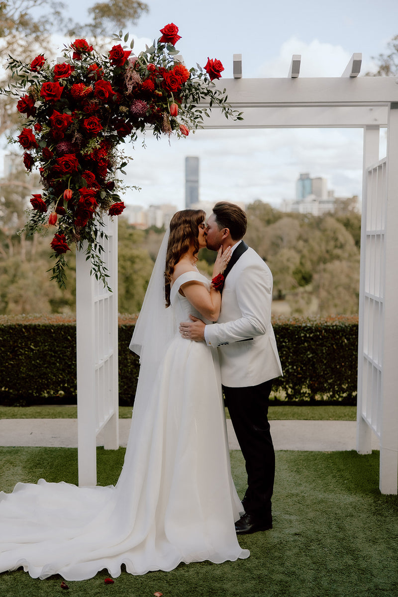 Couple kissing under a red floral arch with a Brisbane cityscape in the background