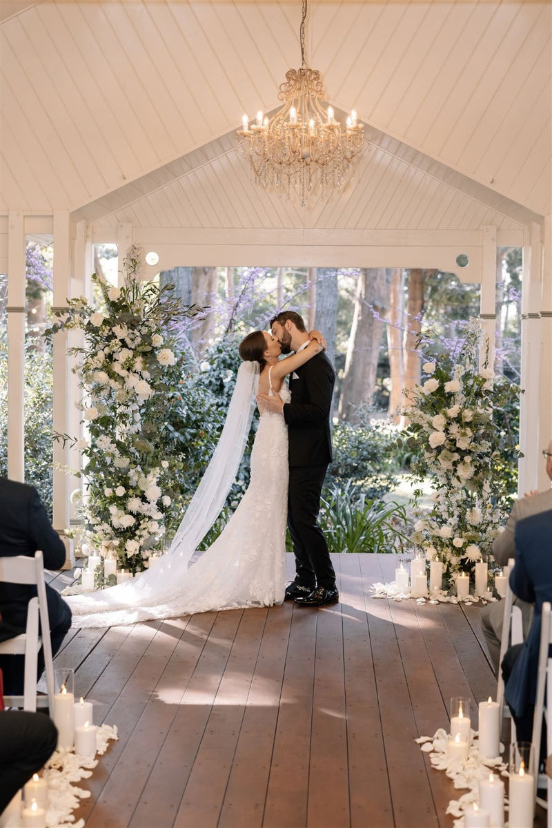 Couple kissing under a chandelier in a decorated indoor setting with floral arrangements.