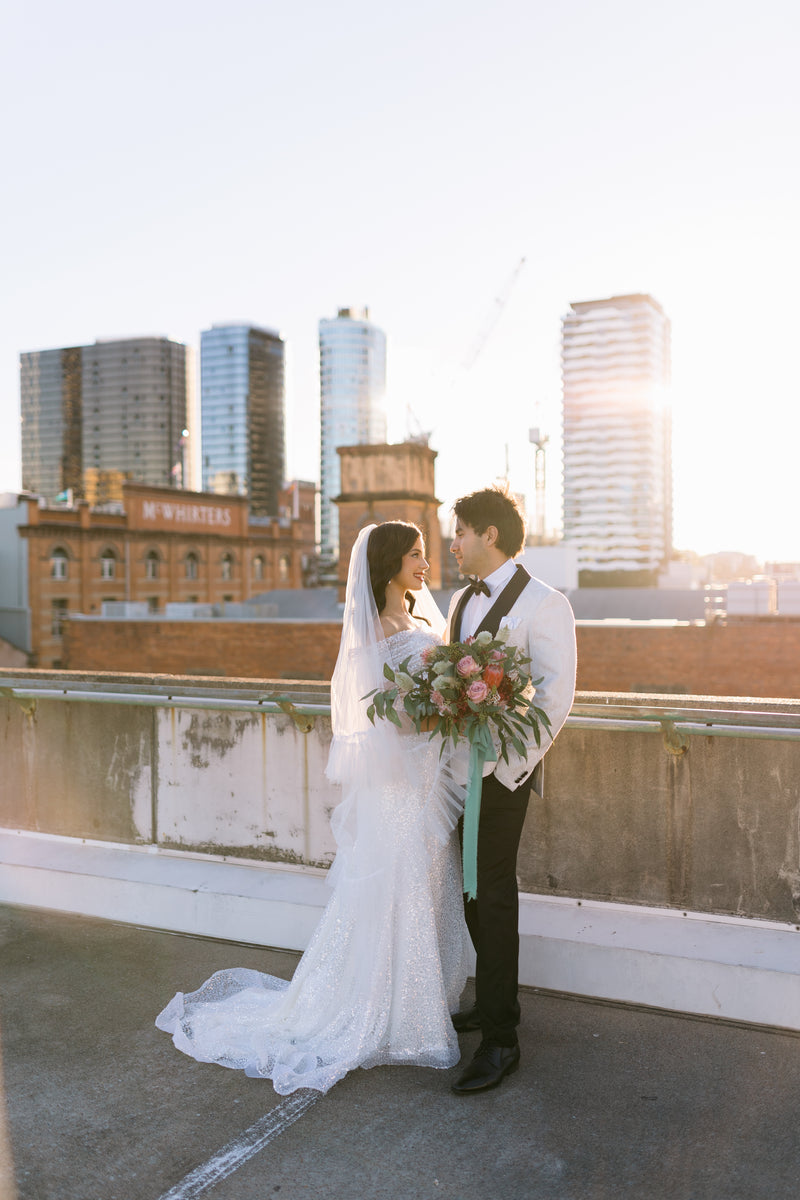 Wedding couple on a rooftop with Brisbane city skyline in the background. Native brides bouquet with a long veil.