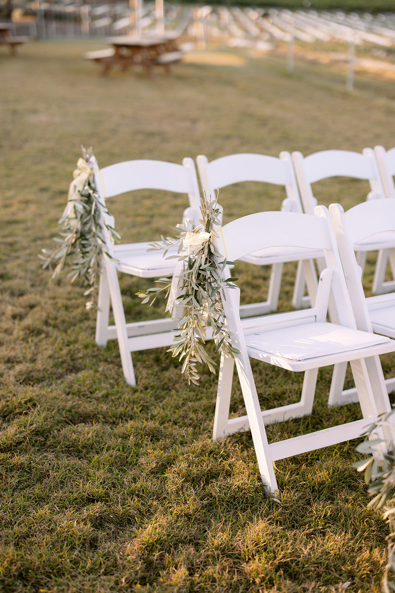 White chairs decorated with olive foliage on a grassy outdoor setting at Wellington Point Farmhouse wedding.