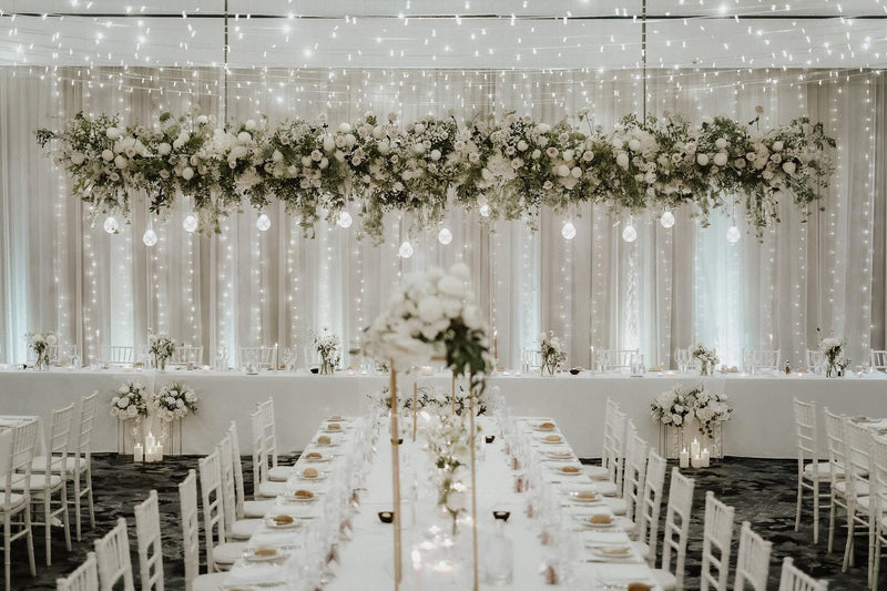 Decorative floral arrangement and string lights hanging above a long table set for a wedding in Byron Bay.